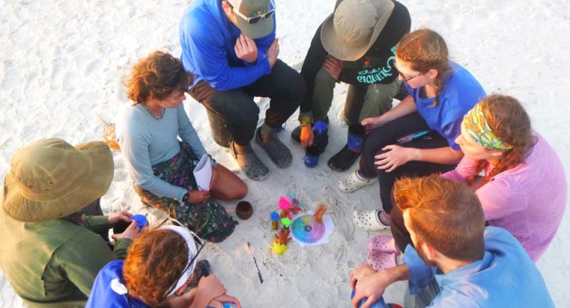 A group of people sit in a circle on white sand. They appear to be looking at something colorful in the center. 